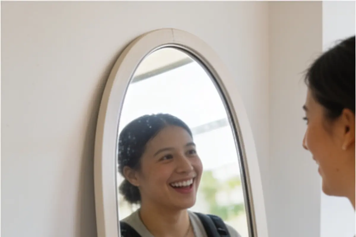 A parent and a baby in a carrier smile at their reflection in a tall mirror.