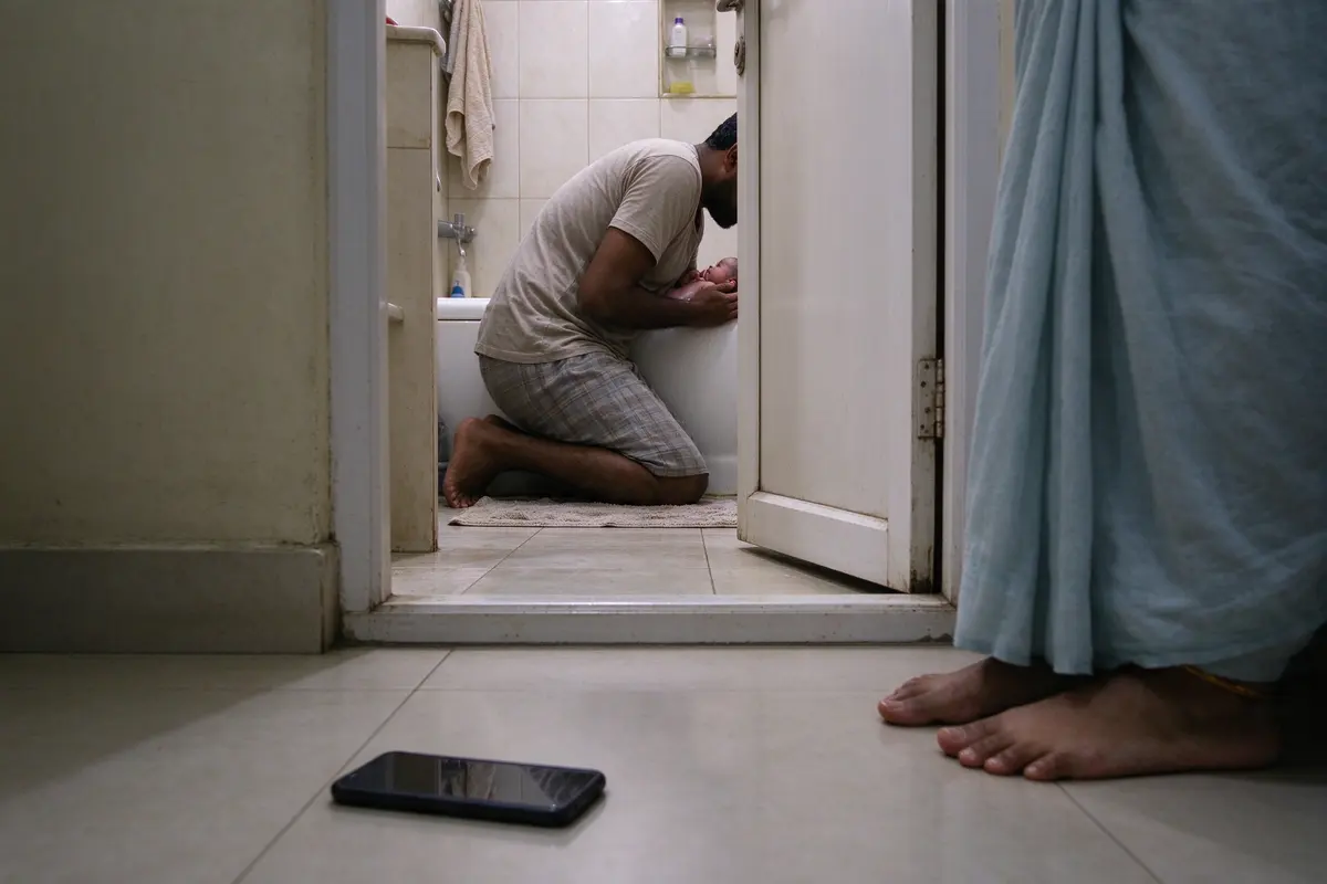 The mother's phone lying face-down on the hallway floor outside the open bathroom door, her bare feet beside it, the father's voice-level view of the bath glimpsed through the doorway.
