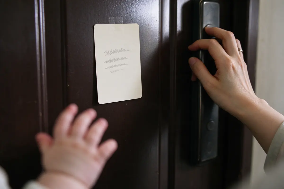Close on an index card taped inside the front door at adult eye level with three handwritten lines, the mother's hand on the door handle beside it, the baby's reaching hand blurred in the foreground.