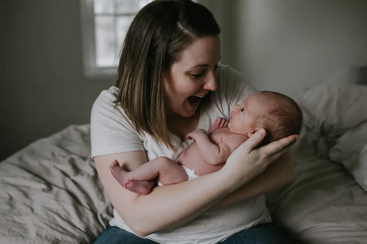 On the edge of the unmade bed in gray morning light the mother holds her two-week-old ten inches from her face, making a slow wide-mouthed expression, the baby's eyes fixed on her.