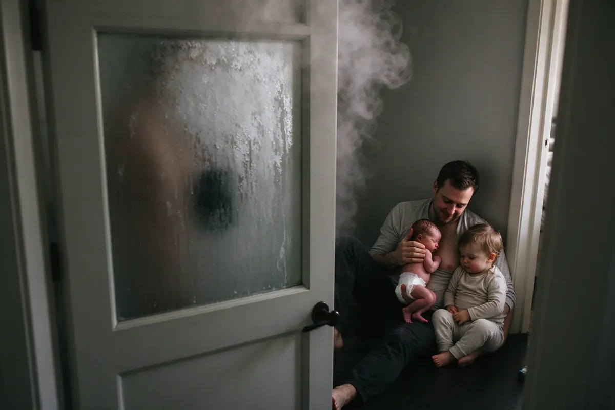 A frosted bathroom door shut, steam curling over the top, while in the hall her partner sits on the floor in soft daylight holding the newborn upright on his chest, the toddler leaning against him.