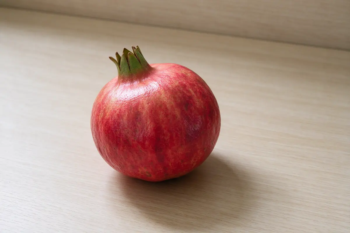 A whole red pomegranate on a plain pale wooden board, soft window light.