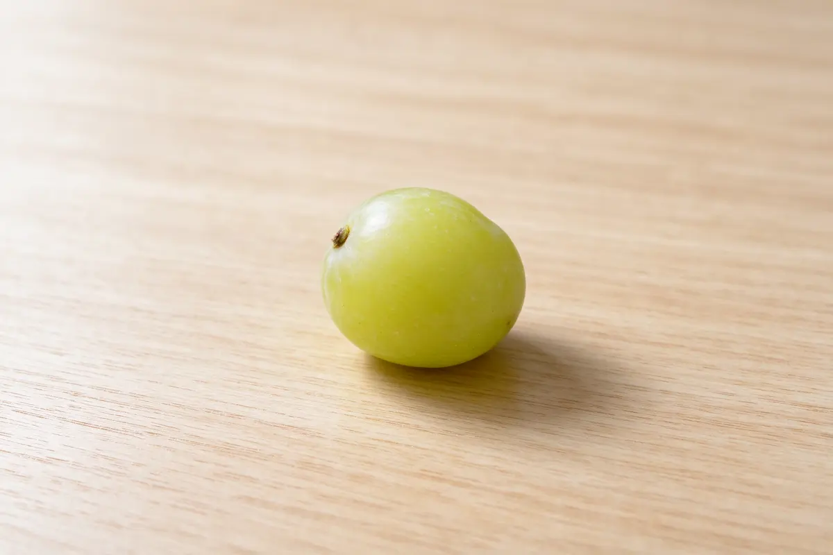 A single green grape on a plain pale wooden table, close up, soft window light.