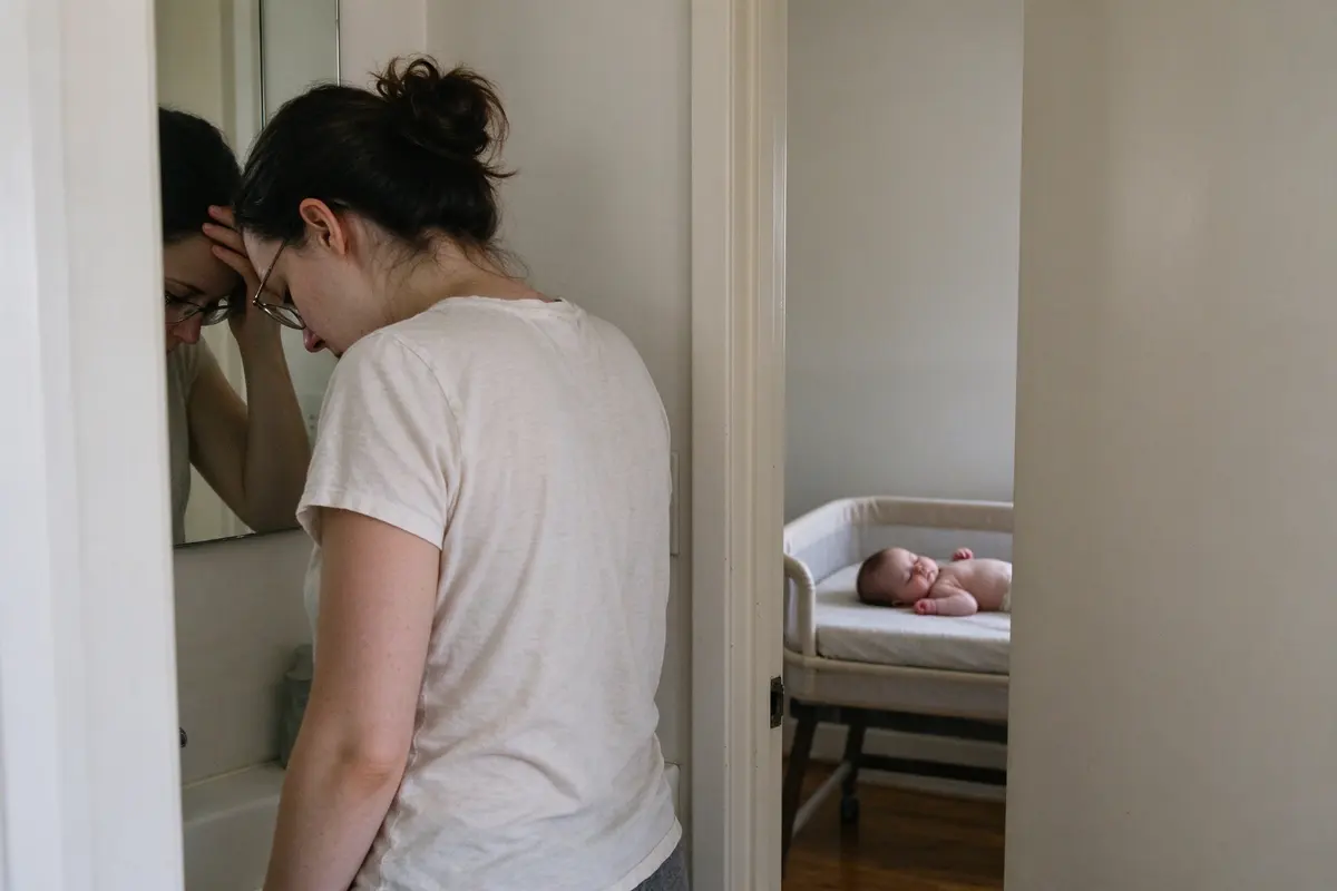 The mother stands at a bathroom mirror in flat daylight with one hand on her forehead, the baby asleep on their back in a bare bassinet through the doorway.