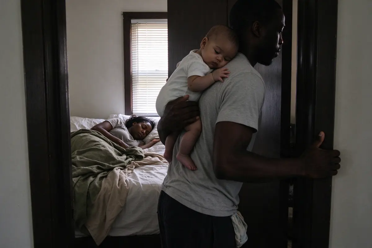 The mother asleep on her side in bed in early-morning light, the father carrying the baby out of the room on his shoulder and pulling the door nearly closed behind him.