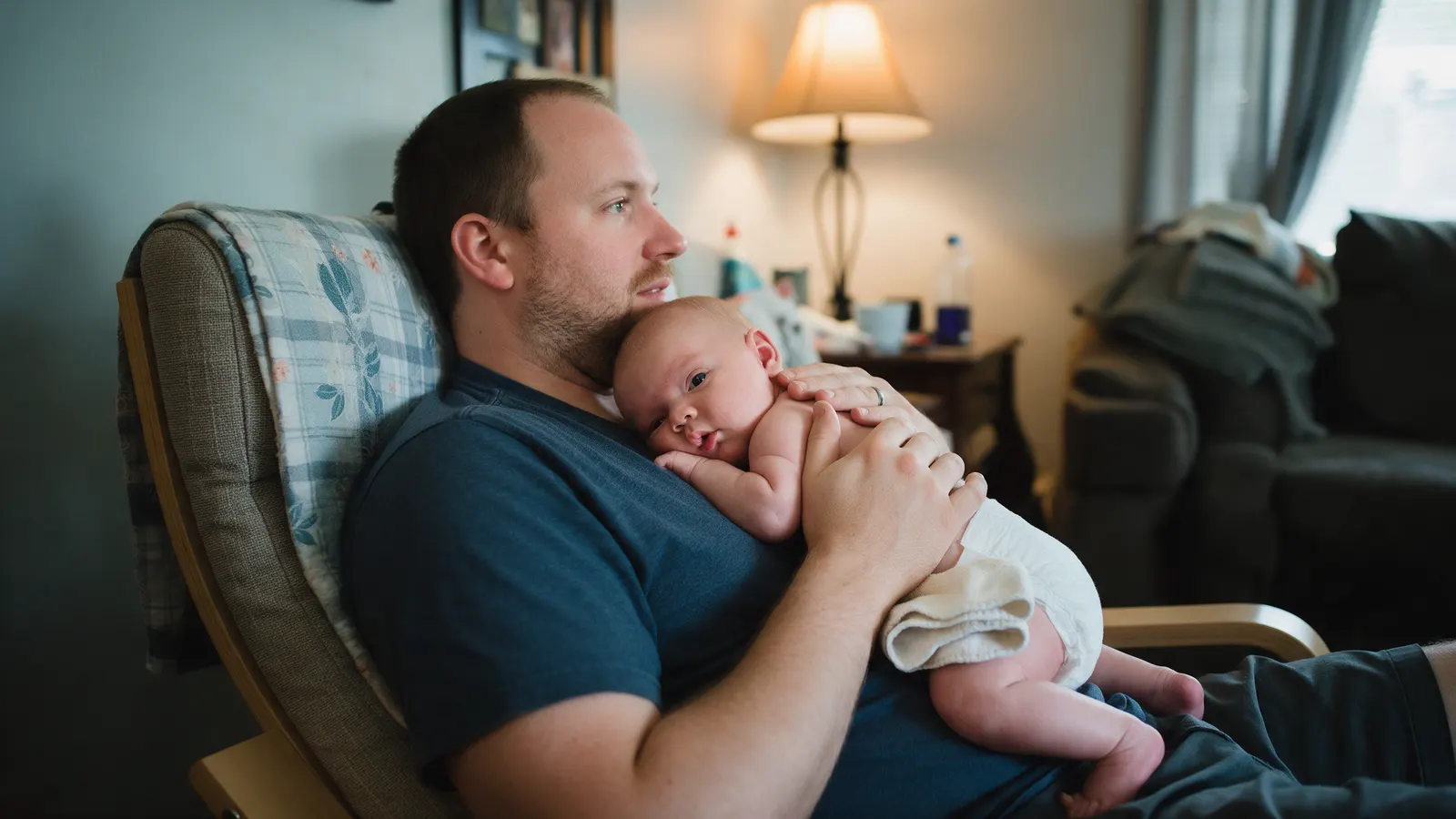 A white father in an armchair holds his nine-week-old against his chest, a folded warm washcloth resting on the baby's thigh, soft lamplight.